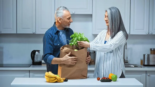 Elderly Interracial Couple Unpacking Paper Bag Fresh Vegetables Fruits Kitchen — Stock Photo, Image