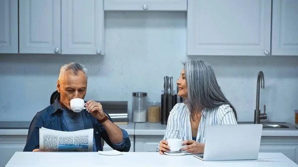 アジア系の女性が高齢者の夫にコーヒーを飲みながらキッチンで新聞を読むのを見て — ストック写真