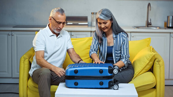 senior interracial couple sitting on couch at home near suitcase
