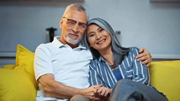 Cheerful Elderly Interracial Couple Smiling Camera While Hugging Sofa Home — Stock Photo, Image