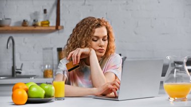 Mutfakta online alışveriş yaparken elinde kredi kartı tutan kıvırcık, genç bir kadın. 