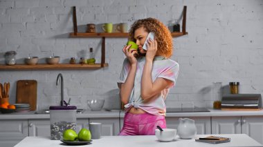Akıllı telefondan konuşan kıvırcık saçlı kadın ve masadaki mısır gevreğinin yanında elma kokusu alıyorum.