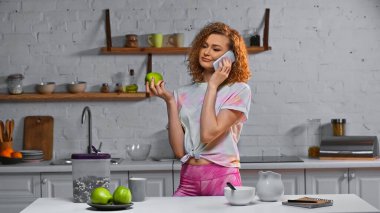 Kıvırcık genç bir kadın akıllı telefondan konuşuyor ve elinde mısır gevreğinin yanındaki masada elma tutuyor.