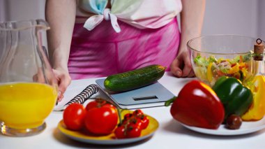 cropped view of woman writing in notebook while weighing cucumber clipart