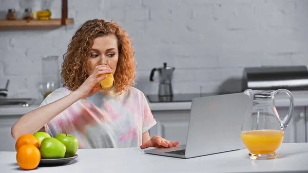 Ricci Giovane Donna Guardando Computer Portatile Mentre Beve Succo Arancia — Foto Stock