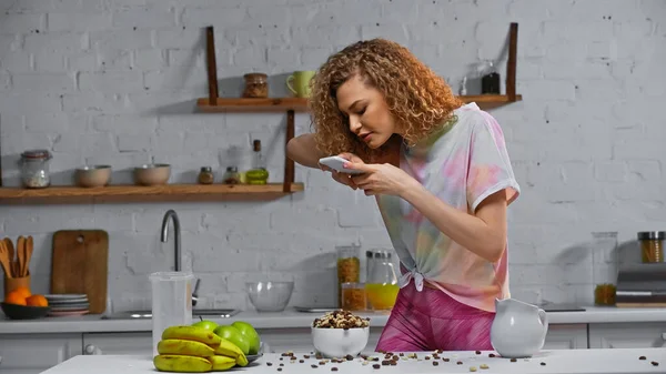 卷曲的年轻女子在厨房桌上给玉米片和水果拍照 — 图库照片