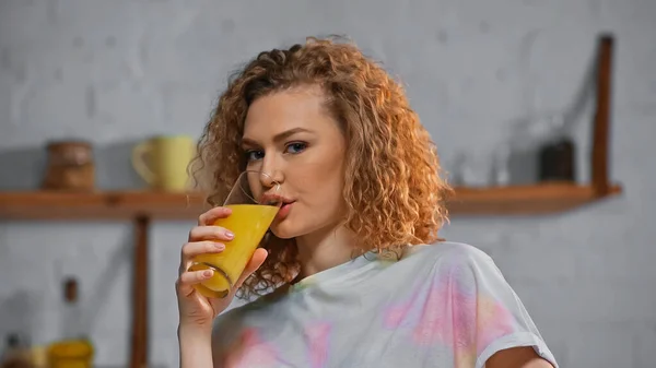 Krøllete Kvinne Som Ser Kamera Mens Hun Drikker Appelsinjuice Kjøkkenet – stockfoto