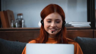 Kulaklık takmış gülümseyen kız bilgisayarında bulanık ön planda görüntülü arama yapıyor. 