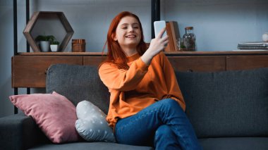 Kanepede yastıkların yanında akıllı telefon kullanan gülümseyen kız. 