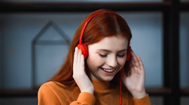 Smiling red haired girl using headphones at home  clipart