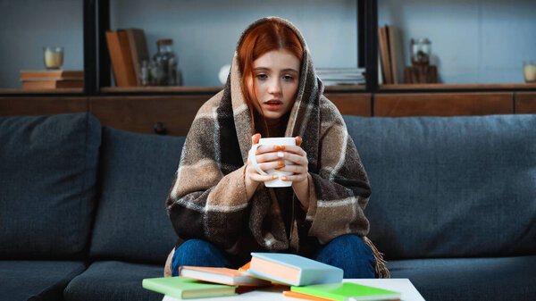 Girl in blanket holding cup near books on blurred foreground 
