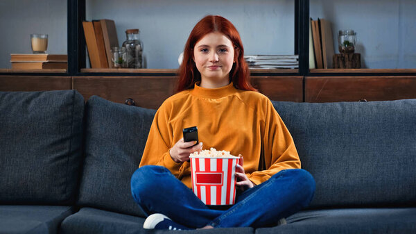 Teenager holding popcorn and remote controller on couch 