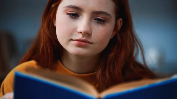 Red haired teenager reading book on blurred foreground at home