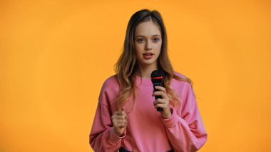 Pembe kazaklı genç kız sarıda izole edilmiş mikrofonu tutarken konuşuyor.
