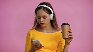 Kablosuz kulaklıklı genç kız akıllı telefon kullanıyor ve pembe bardakta izole edilmiş kağıt bardak tutuyor. 