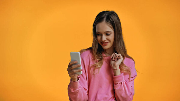 cheerful teenage girl in pink sweatshirt looking at smartphone isolated on yellow
