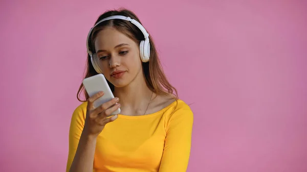 teenage girl in wireless headphones using smartphone isolated on pink