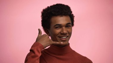 happy african american teenage boy in turtleneck sweater showing call me gesture isolated on pink  clipart