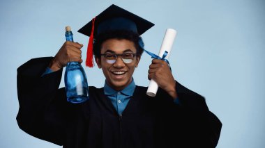 Mezuniyet elbisesi ve şapkalı Afro-Amerikan genç elinde diploma ve şişe ile mavi üzerine izole edilmiş şampanya tutuyor.