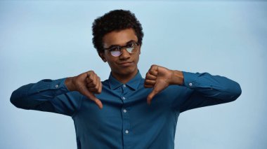 african american teenage boy in glasses showing thumbs down isolated on blue clipart