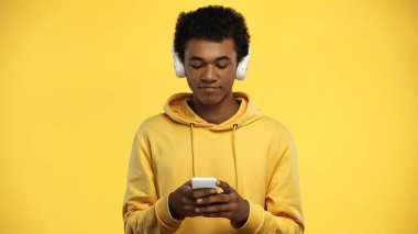 Sarı kablosuz kulaklıklı Afro-Amerikan genç çocuk sarıda izole edilmiş akıllı telefon kullanıyor.