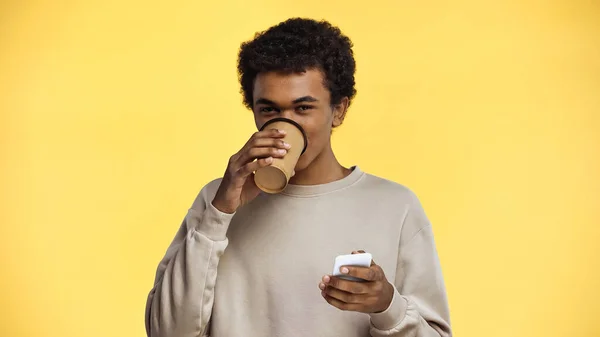 アフリカ系アメリカ人の10代の若者がコーヒーを飲みながら黄色に隔離されたスマートフォンを持ち — ストック写真