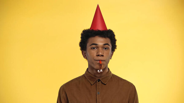 african american teenager in party cap blowing horn isolated on yellow