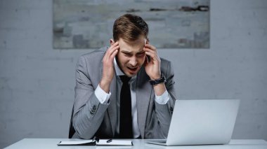 upset businessman in suit touching head while having migraine in office