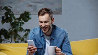 happy man holding smartphone and cup of coffee in living room 