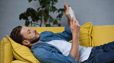 bearded man lying on sofa and reading book in living room