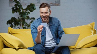happy man holding credit card near laptop while sitting on sofa
