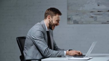side view of bearded businessman laughing while using laptop in office