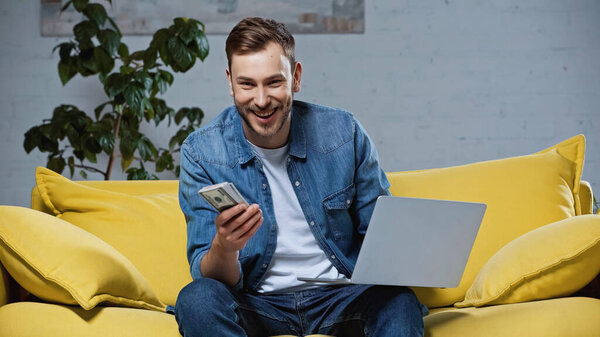 счастливый человек, держащий доллары рядом с ноутбуком, сидя на диване