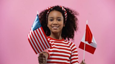 happy african american girl holding flags of america and canada isolated in pink 