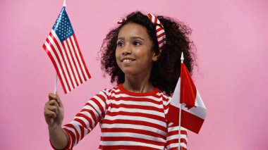 smiling african american girl holding flags of america and canada isolated in pink 