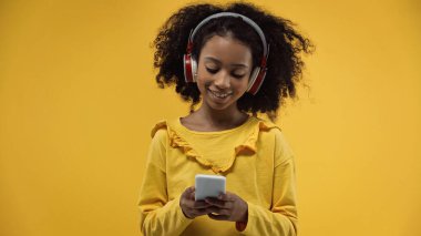 curly african american girl in headphones using smartphone isolated on yellow
