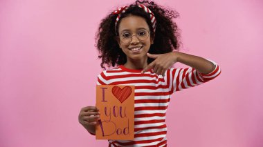 happy african american girl in glasses pointing with finger at greeting card with i love you dad lettering isolated on pink