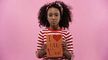 curly african american girl in glasses holding greeting card with i love you dad lettering isolated on pink