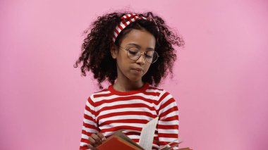 curly african american girl in glasses reading novel isolated on pink 
