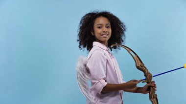 happy african american girl with wings holding crossbow isolated on blue