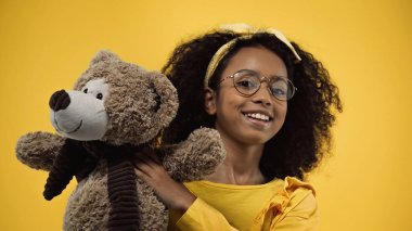happy african american girl in glasses holding teddy bear isolated on yellow clipart