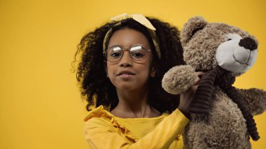 curly african american kid in glasses holding teddy bear isolated on yellow clipart