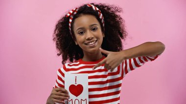 happy african american girl pointing with finger at greeting card with i love mom lettering isolated on pink