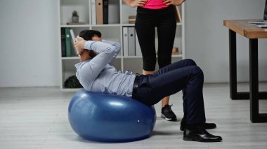 Fitness trainer standing near businessman training on fitness ball in office  clipart