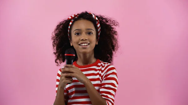 cheerful african american girl holding microphone isolated on pink