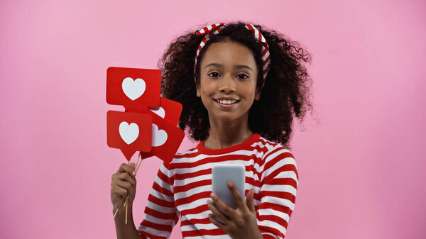 happy african american girl holding smartphone and hearts on sticks as likes isolated on pink