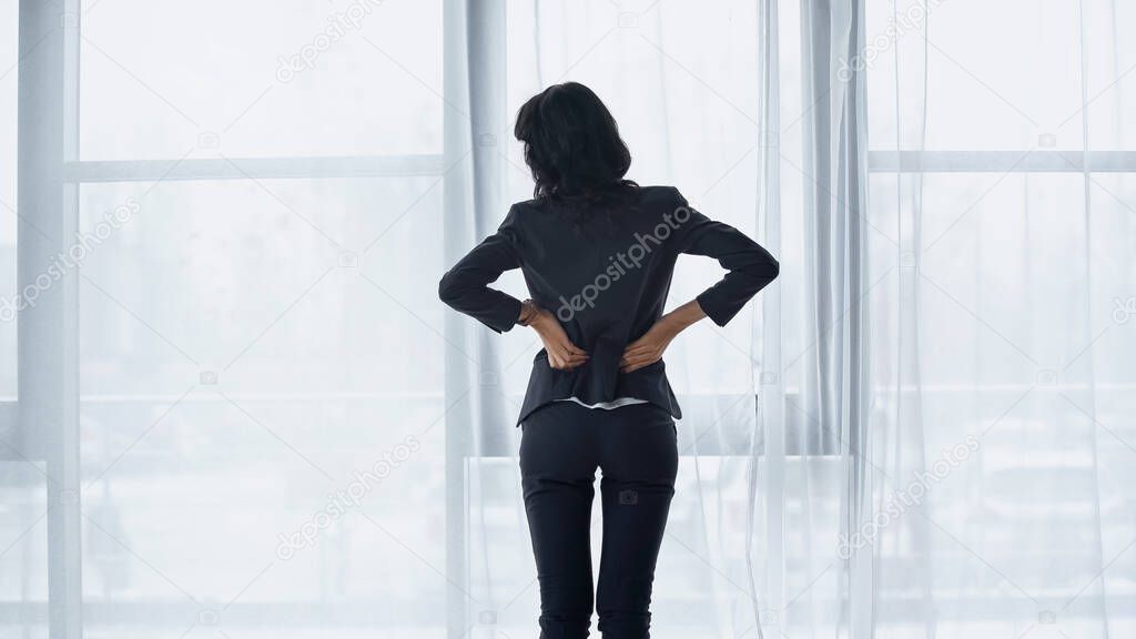 Back view of businesswoman feeling back pain while standing near window in office 