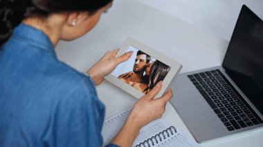 Genç bir kadın fotoğraf çerçevesini dizüstü bilgisayarın yanında tutuyor.