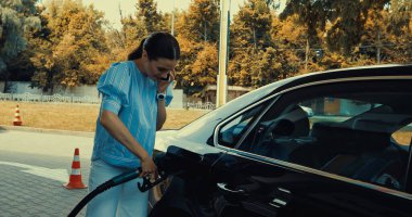 Benzin istasyonunda yakıt ikmali yaparken akıllı telefondan konuşan genç bir kadın.
