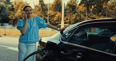 Benzin istasyonunda arabaya yakıt doldururken akıllı telefondan konuşan mutlu bir kadın.